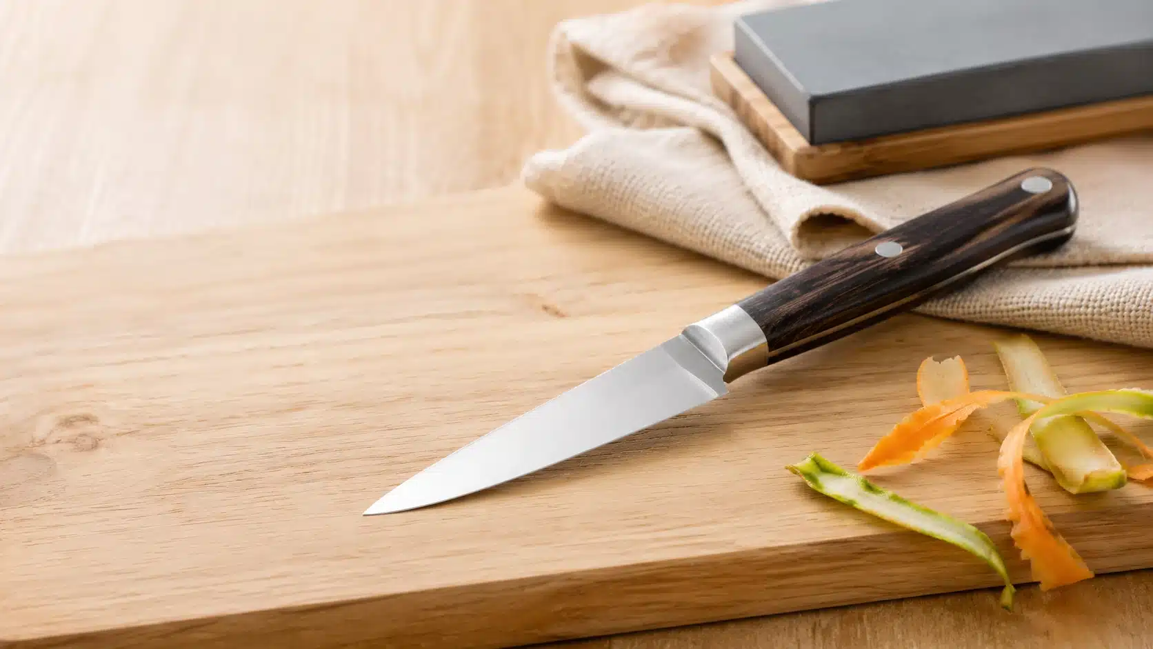 Un couteau d'office au manche en bois sombre posé sur une planche de découpe en bois d'un beige clair et lumineux, à côté de fines épluchures de légumes avec une pierre à aiguiser sur un linge en arrière-plan.