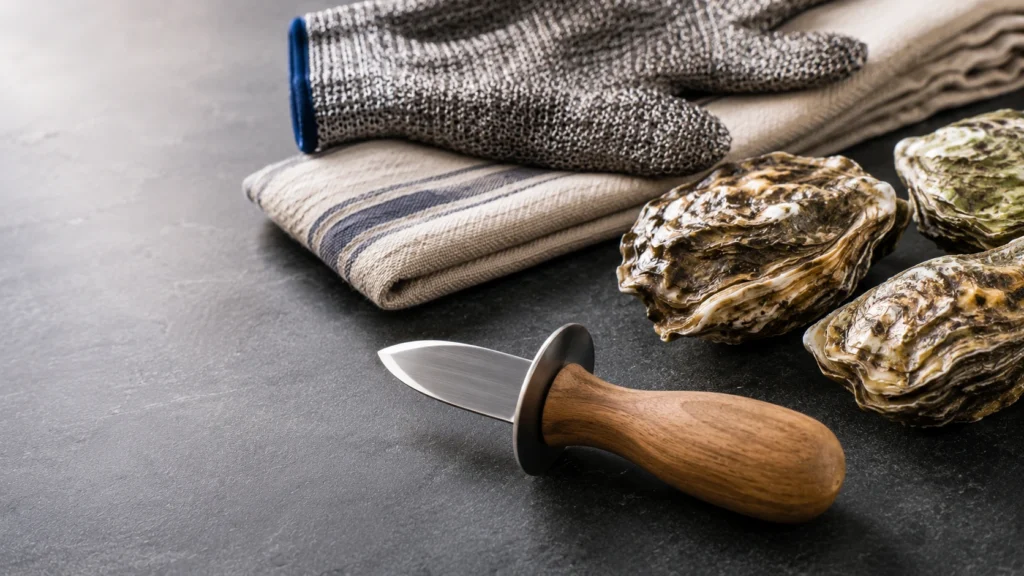 Une vue en plongée d'un couteau à huître ergonomique avec un manche en bois et une garde, prêt à l'emploi. Il est posé sur un plan de travail en ardoise sombre, à côté de trois huîtres fraîches, d'un gant de protection en maille et de deux serviettes en tissu.