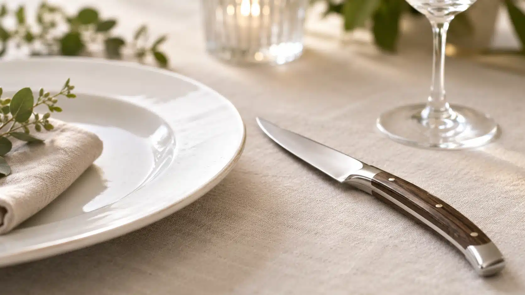 Un couteau de table artisanal au design courbé et élégant, doté d'une lame en acier inoxydable brillante et d'un manche en bois sombre veiné avec rivets en laiton. Le couteau est délicatement posé sur une nappe en lin beige clair, à côté d'une assiette en céramique blanche texturée, d'une serviette et du pied d'un verre à vin.