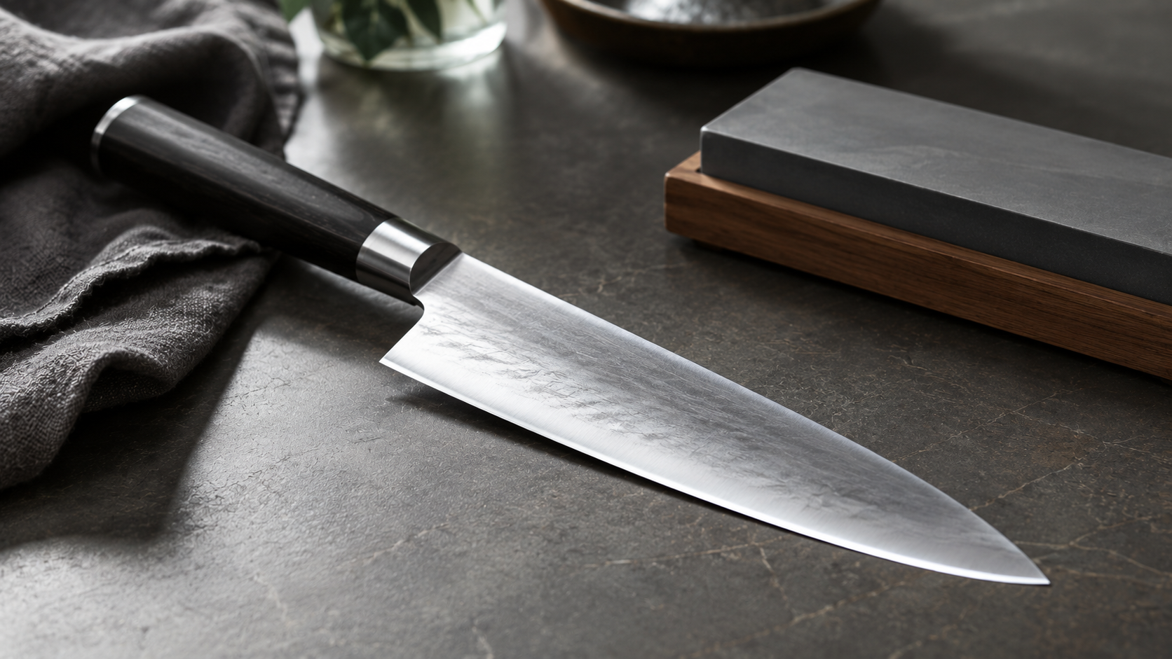 Un couteau en acier carbone de type chef avec une lame brute et un manche en bois noir, disposé à côté d'une pierre à aiguiser traditionnelle sur un plan de travail sombre.